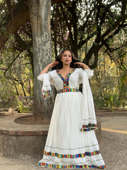 Woman in a white Habesha Kemis with colorful embroidery standing outdoors near trees.