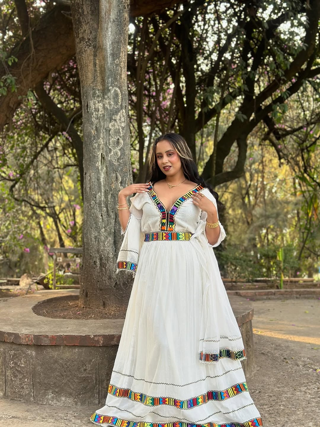 Woman in a white Habesha Kemis with colorful patterns standing outdoors near trees.