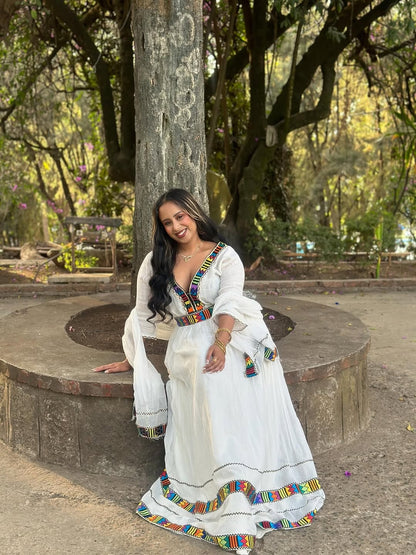 Woman in a white Habesha Kemis with colorful patterns sitting outdoors.