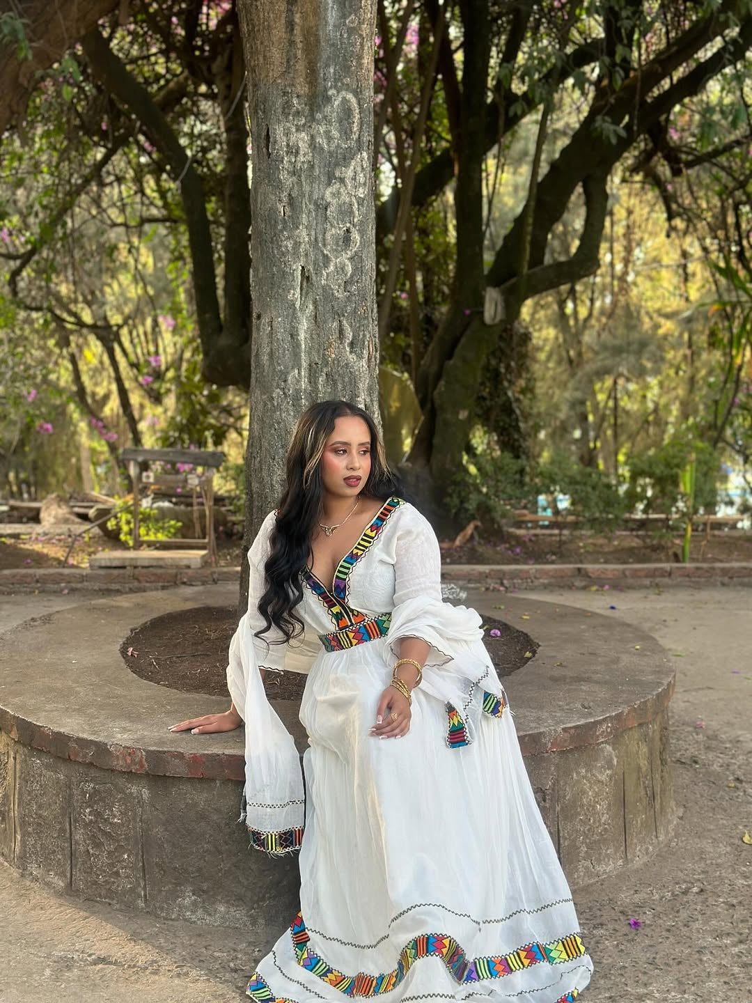 Woman in a white Habesha Kemis with colorful accents sitting on a stone bench in a park.