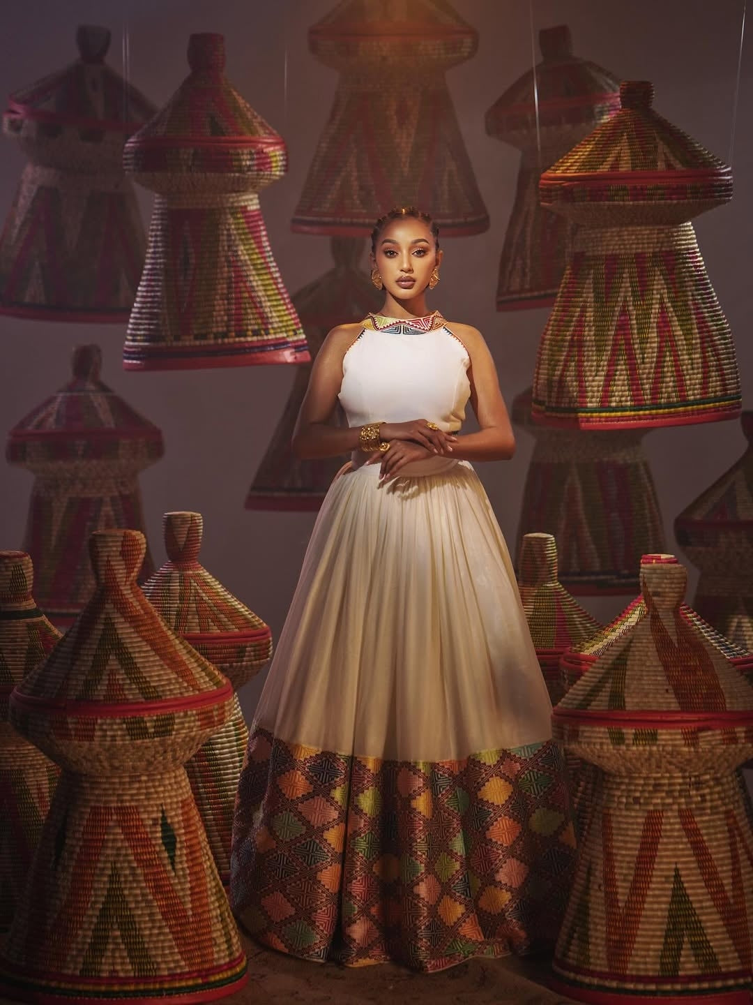 Woman in a white dress standing among traditional woven baskets with geometric patterns.