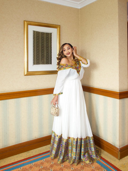 Woman in a white habesha Libs with floral patterns standing in a room with decorative walls.