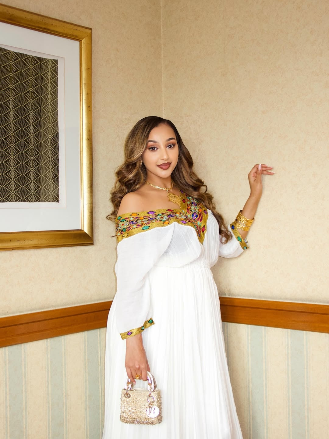 Woman in a white habesha Libs with gold and green embroidery standing against a neutral wall.