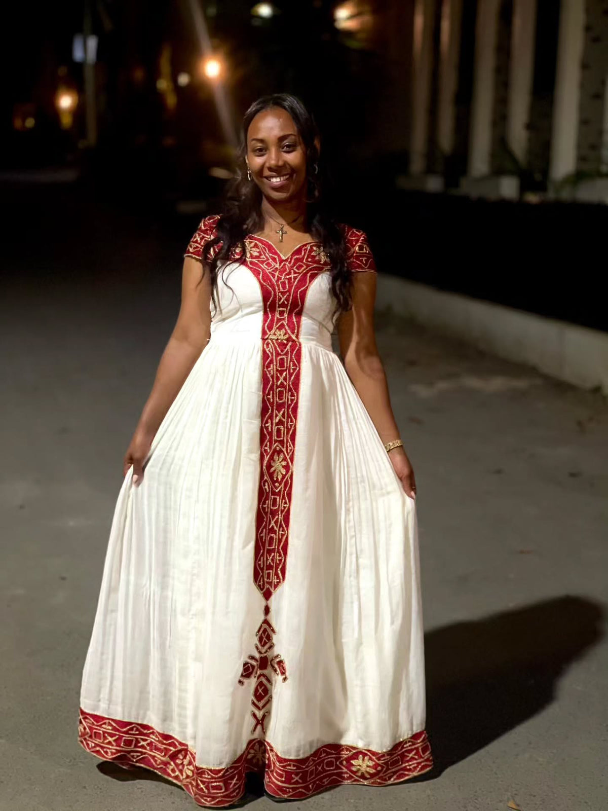 A woman wearing a white Ethiopian Dress with red and gold embroidery, standing outdoors at night on a paved path.