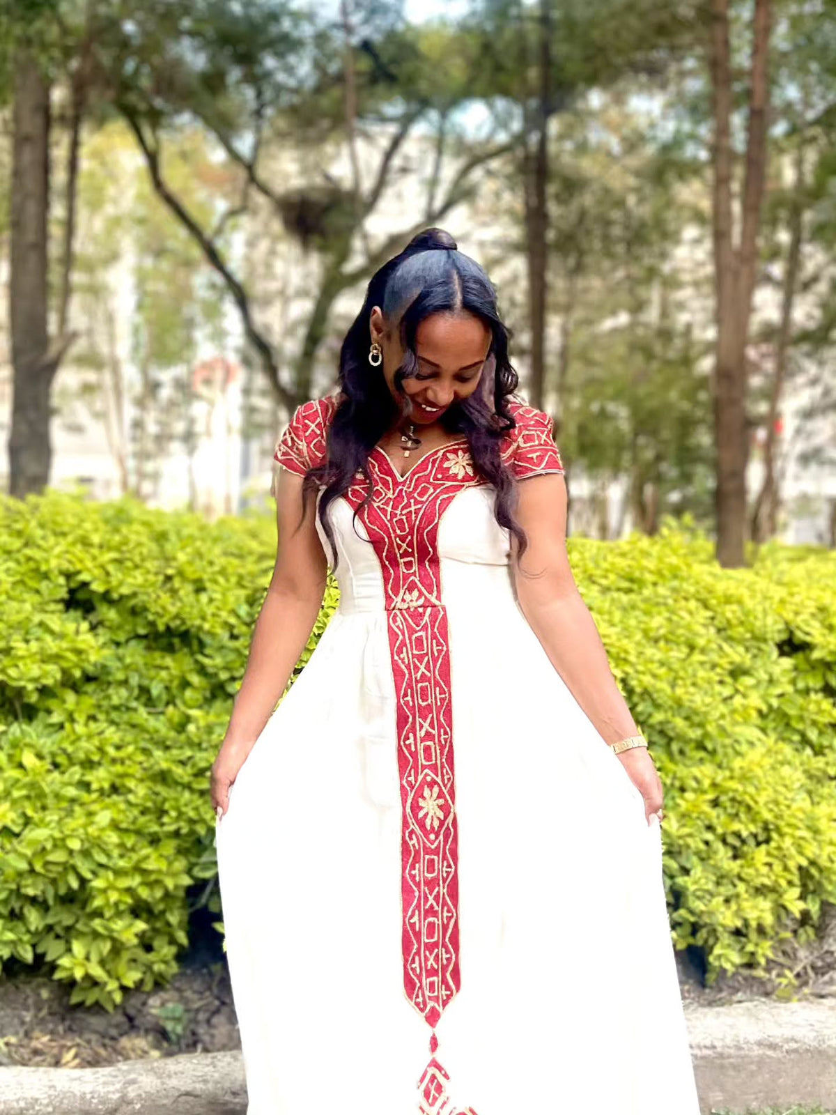 A woman wearing a white Ethiopian Dress with red and gold embroidery, standing outdoors in front of green bushes and trees.