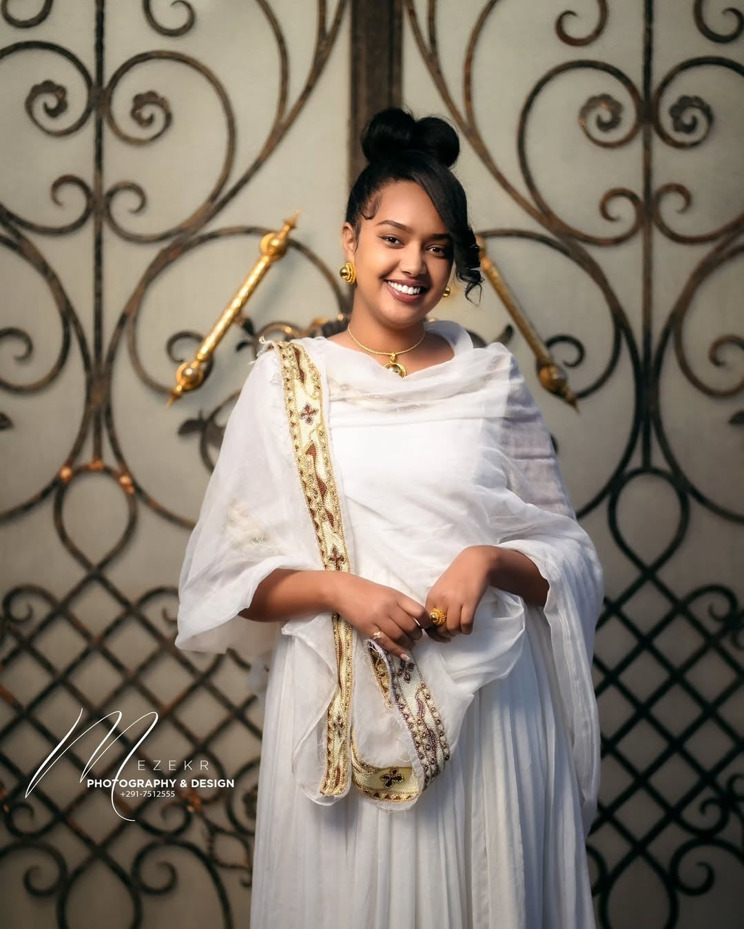 Woman in a white Habesha Zuria  with gold accents standing in front of an ornate metal gate.