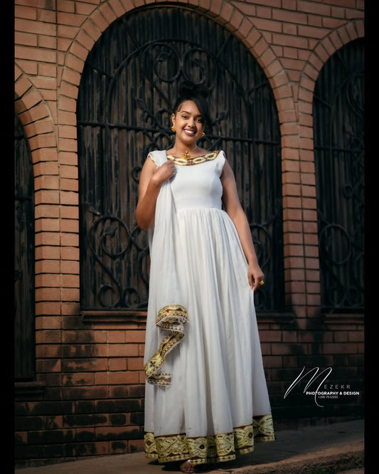 Woman in a white and gold Habesha Zuria standing in front of a brick wall with an arched door.