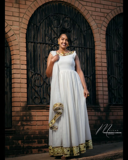 Woman in a white and gold Habesha Zuria  standing in front of a brick wall with an arched door.