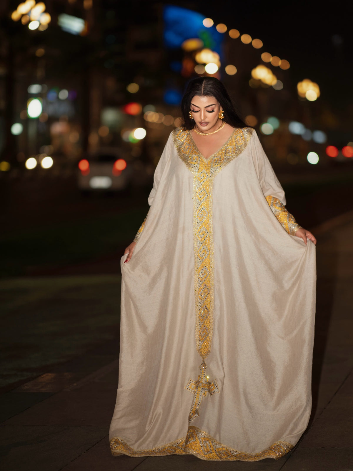 A woman wearing a flowing beige abaya with ornate gold Ethiopian clothing, standing on a city street at night with blurred lights in the background.