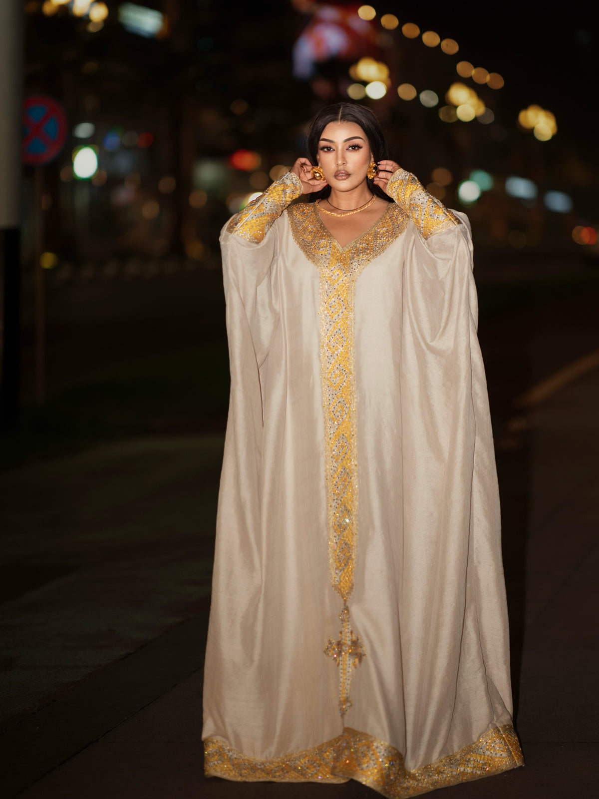 A woman wearing a long beige or cream Ethiopian clothing with ornate gold embroidery and embellishments, standing on a city street at night with blurred lights in the background.