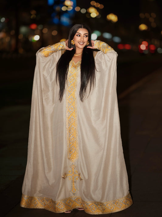 A woman wearing a flowing beige or cream caftan-style Ethiopian clothing with ornate gold embroidery and a gold border, standing outdoors at night with blurred city lights in the background.