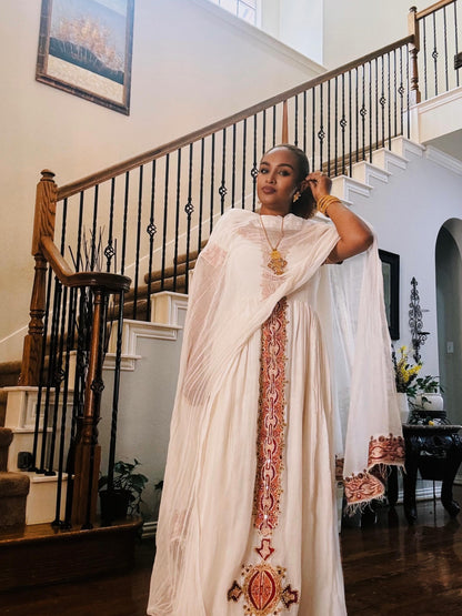 A woman wearing a white traditional Ethiopian traditional clothing with intricate red and gold embroidery, standing in a grand staircase hallway.