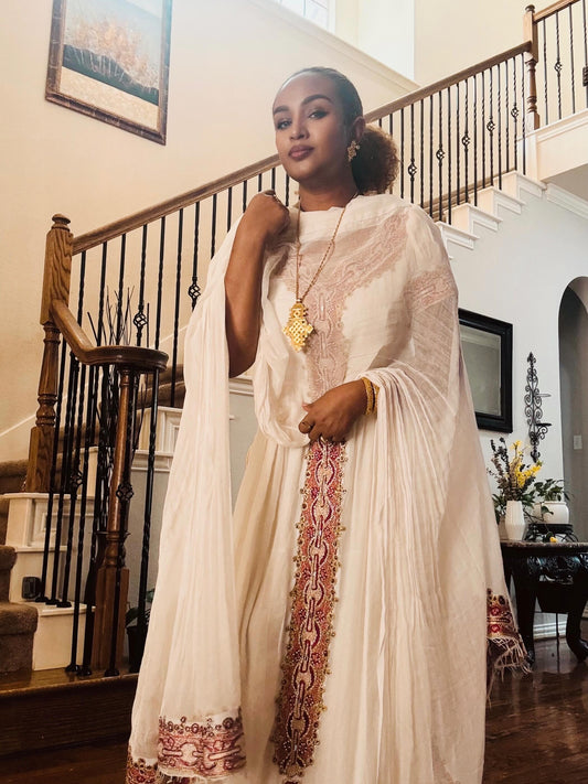 A woman models a traditional cream-colored embroidered Ethiopian traditional clothing with a matching dupatta in an elegant indoor setting with a staircase in the background.