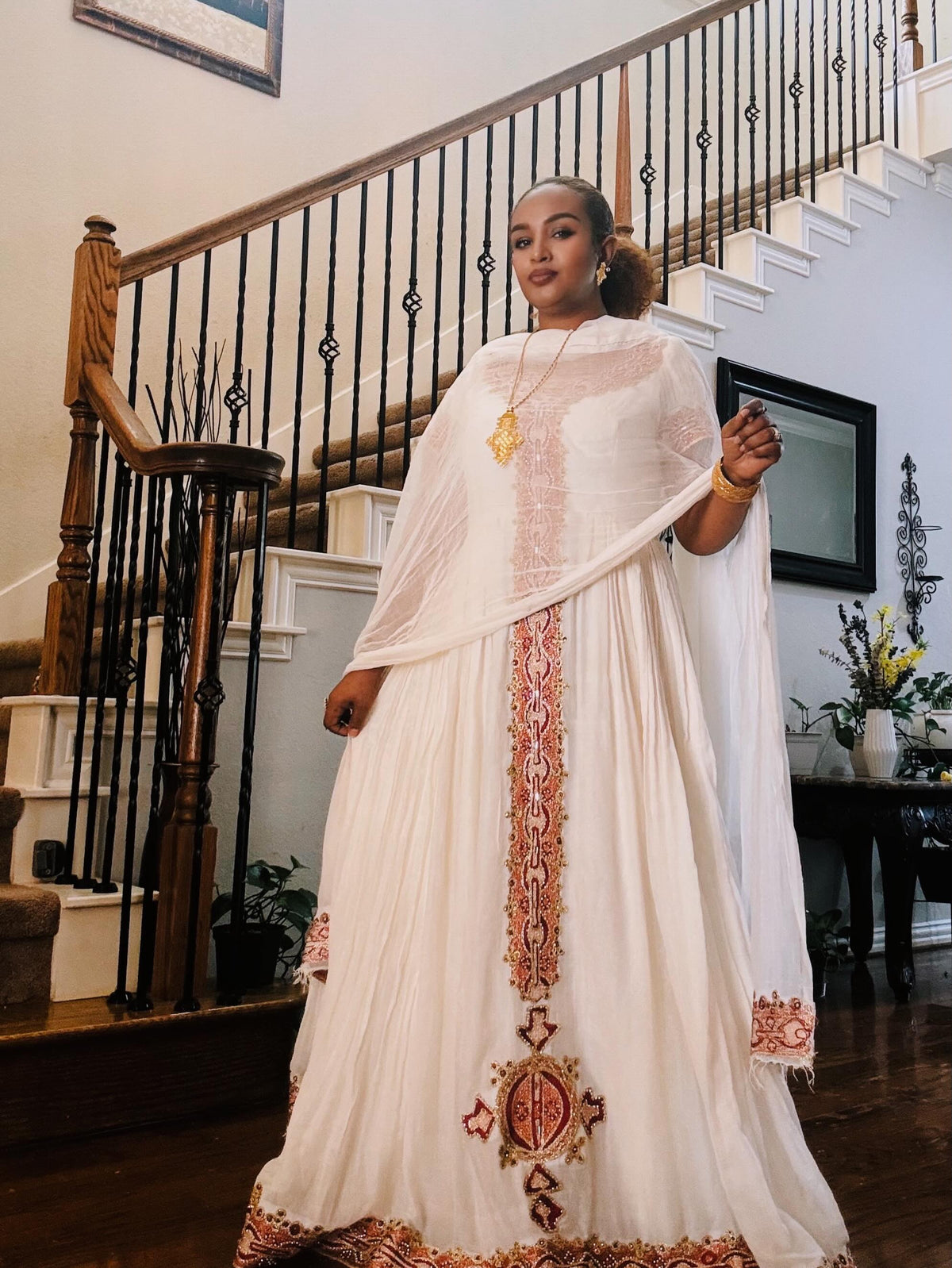 A woman wearing a traditional white embroidered Ethiopian traditional clothing with a matching sheer dupatta and a long embroidered skirt, standing in an indoor setting with a staircase in the background.