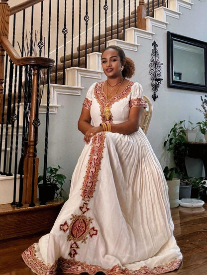 A woman wearing a traditional white Ethiopian traditional clothing with red and gold embroidery, standing in a home interior in front of a staircase.