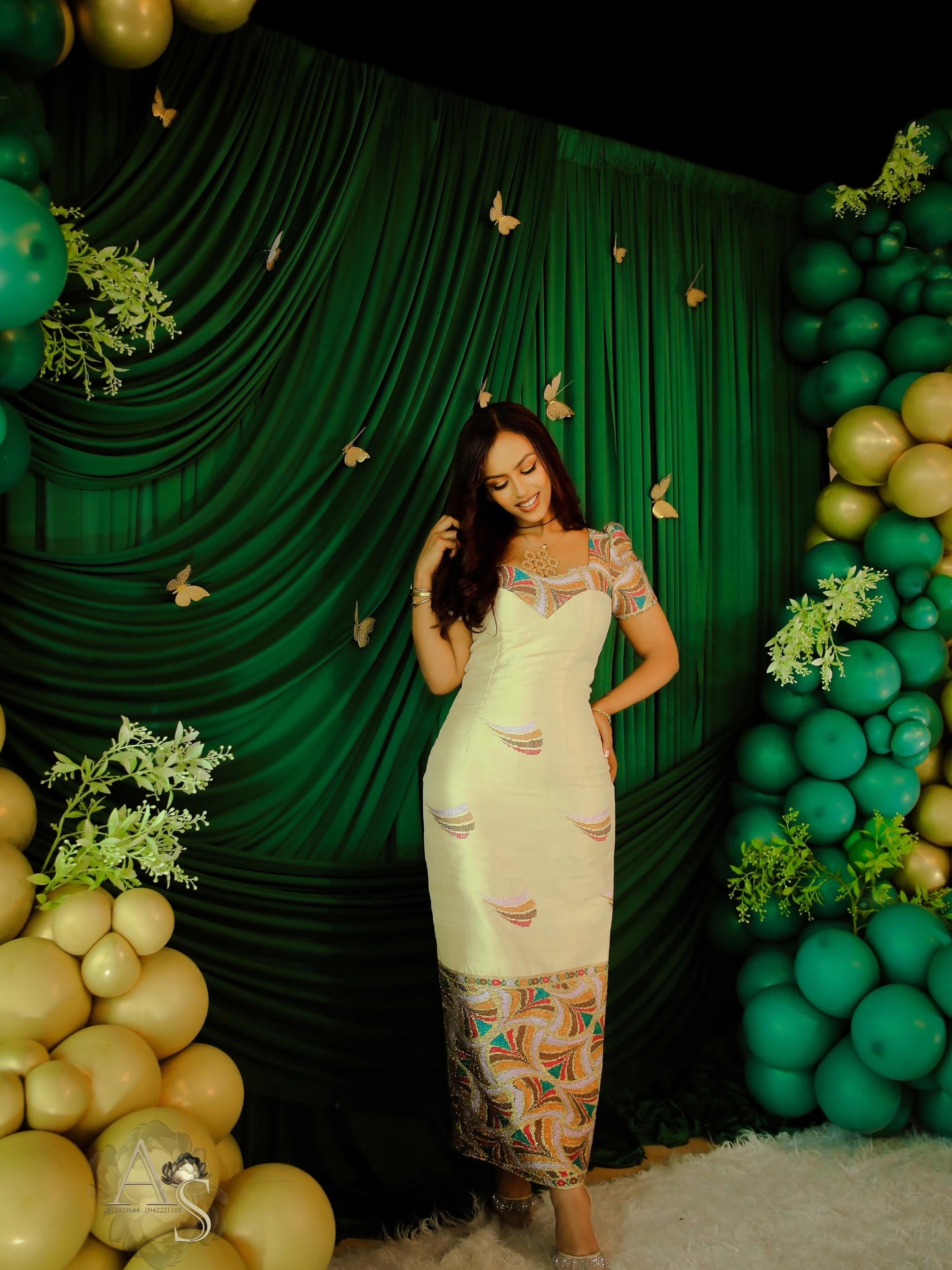 Woman posing in a decorated habesha dress with green and gold balloons and a green curtain.