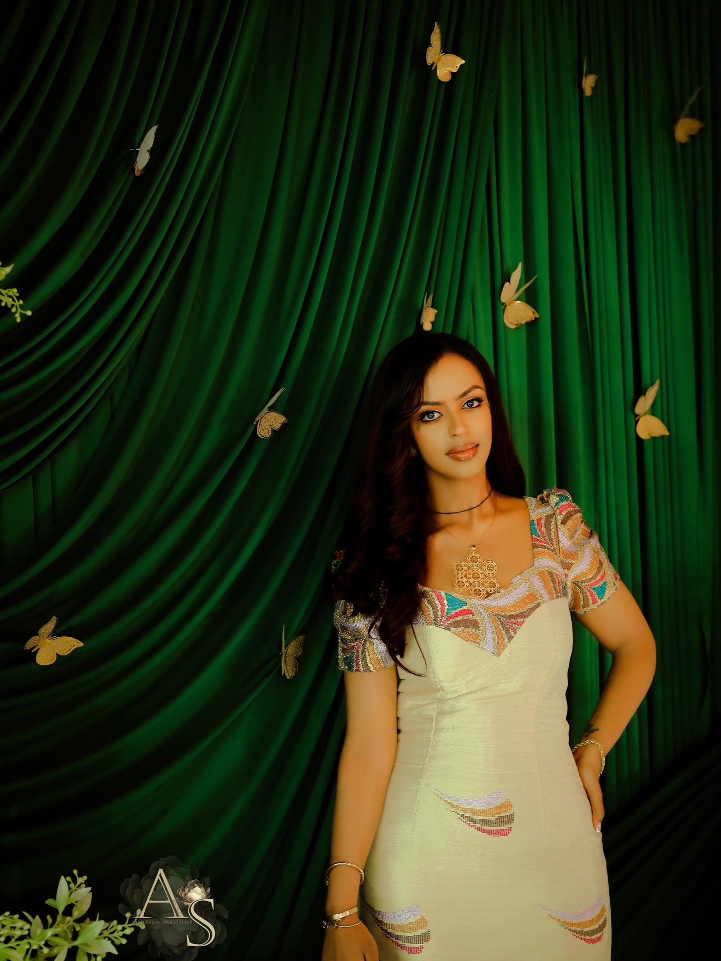 Woman in a white habesha dress with colorful patterns standing in front of a green curtain with butterfly decorations.