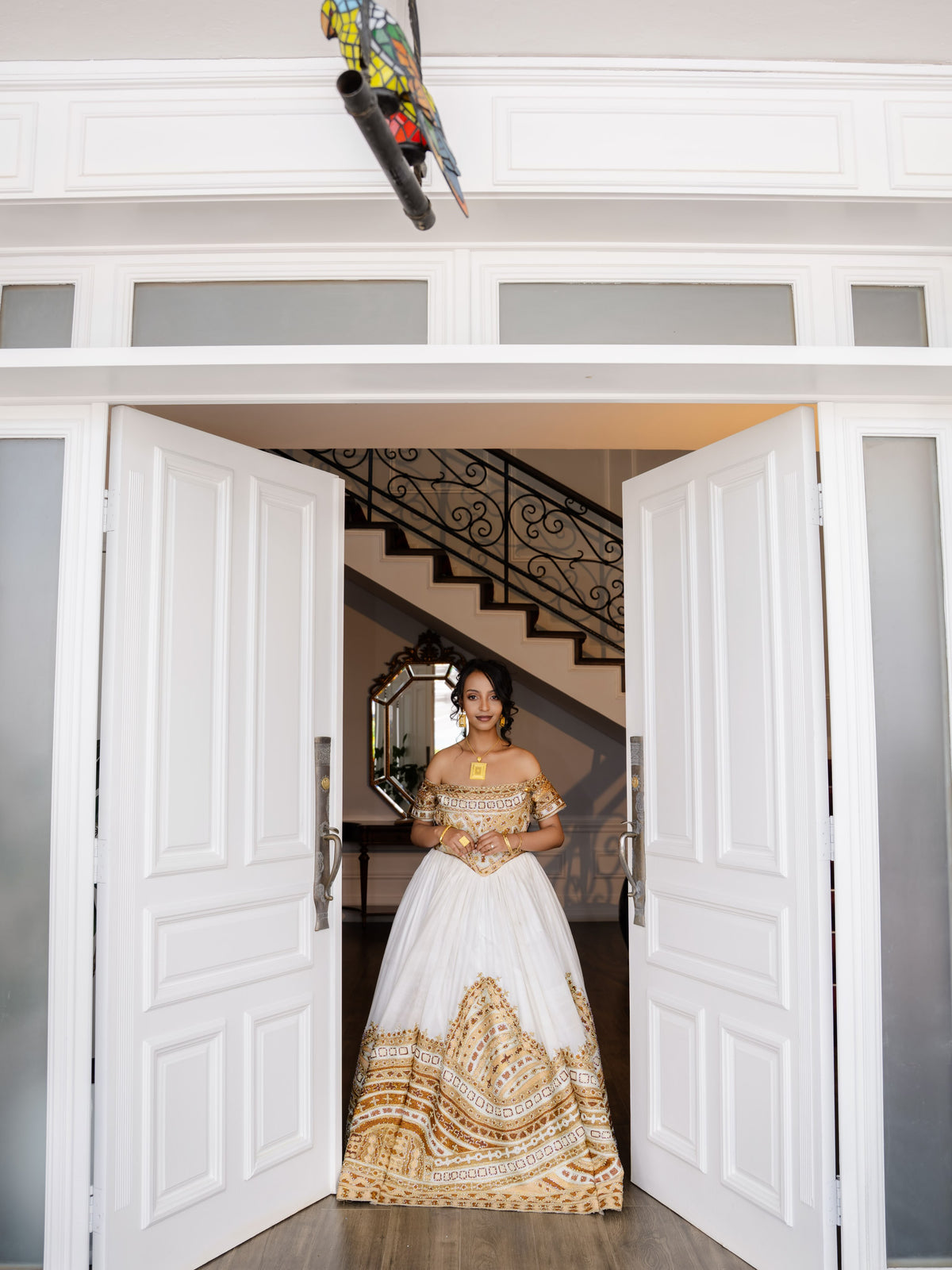 A woman wearing a white ball Ethiopian national dresses with gold embroidery stands in a doorway, looking directly at the camera. The dress features a structured white bodice and a voluminous skirt with intricate gold patterns.