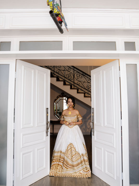 A woman wearing a white ball Ethiopian national dresses with gold embroidery stands in a doorway, looking directly at the camera. The dress features a structured white bodice and a voluminous skirt with intricate gold patterns.
