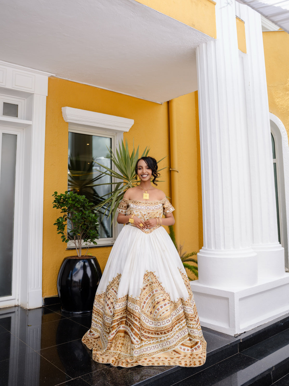 A woman wearing a white ball Ethiopian national dresses with gold and beige bohemian-style patterns, standing on a black tiled porch in front of a yellow and white building.