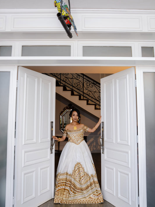 A woman stands in a grand white doorway wearing an off-the-shoulder white Ethiopian national dresses with extensive gold embroidery concentrated on the bodice and a large gold patterned hem. She is holding open the doors, with a staircase visible in the background.