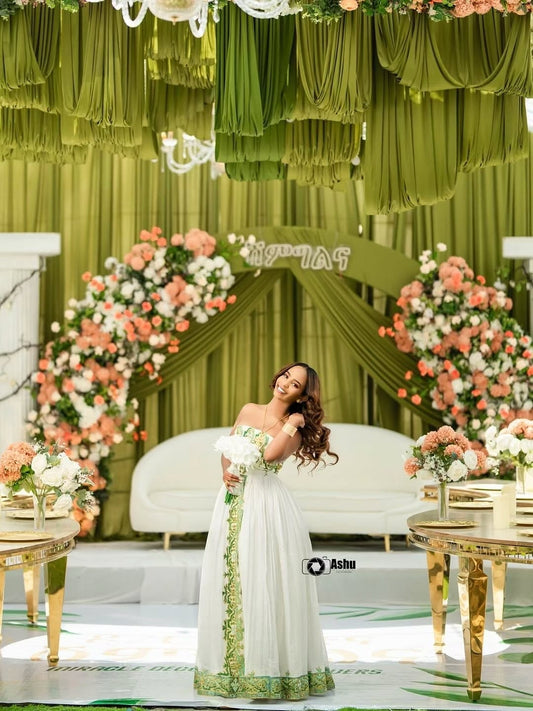 Woman in a white  Habesha Bridal Dress standing in front of a decorated green and white stage with floral arrangements.
