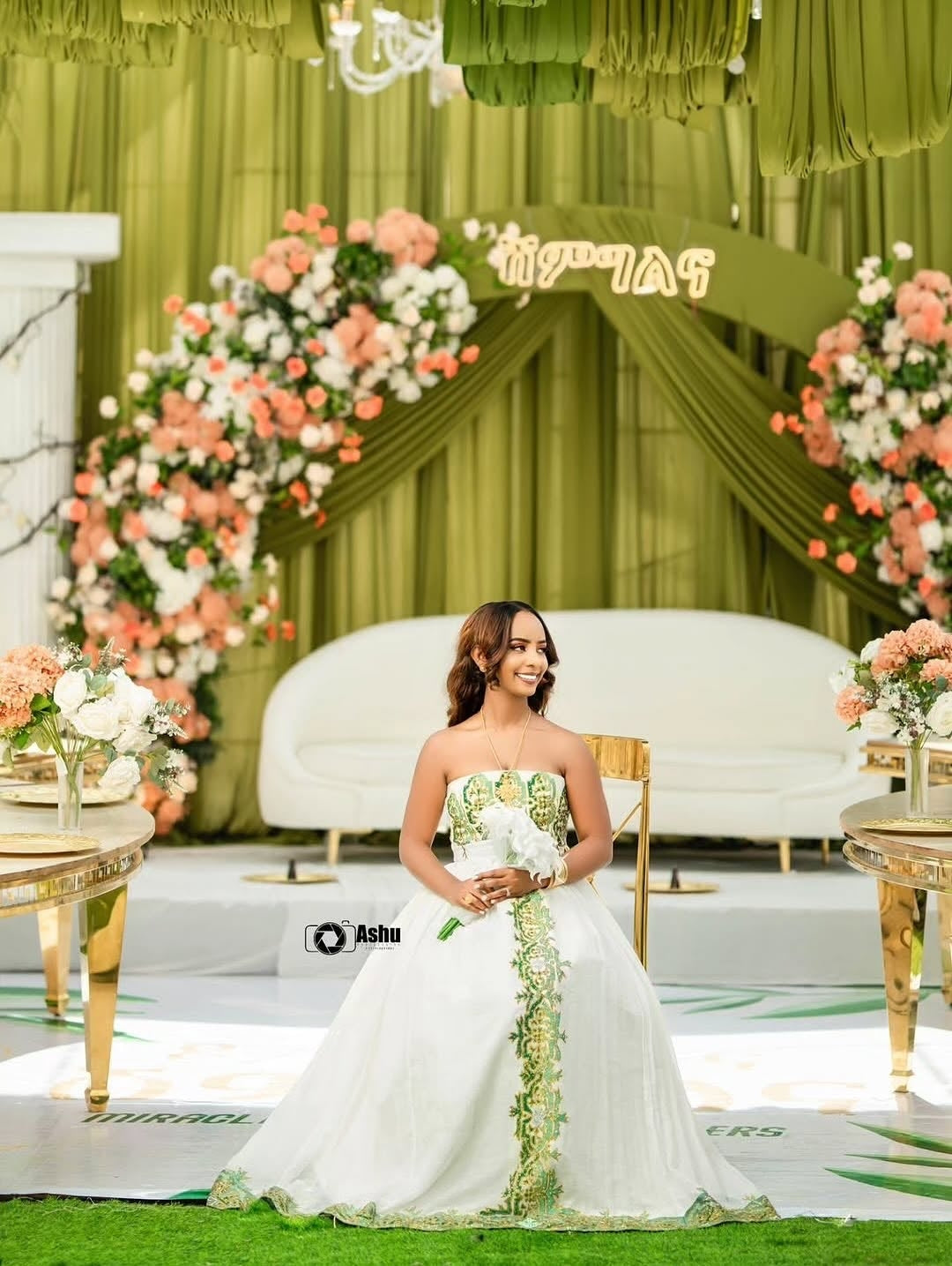 Woman in a white and green  Habesha Bridal Dress standing in front of a decorated stage with floral arrangements and green curtains.