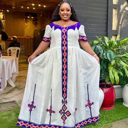 A woman wearing a white A-line Habesha Dress with purple and pink geometric embroidery, holding out the sides of the skirt to show its full length in an outdoor event setting.