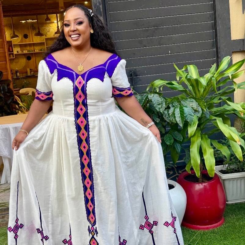 A woman wearing a white African-style Habesha Dress with purple and pink geometric embroidery, holding out the skirt to show its full length in an outdoor patio setting.