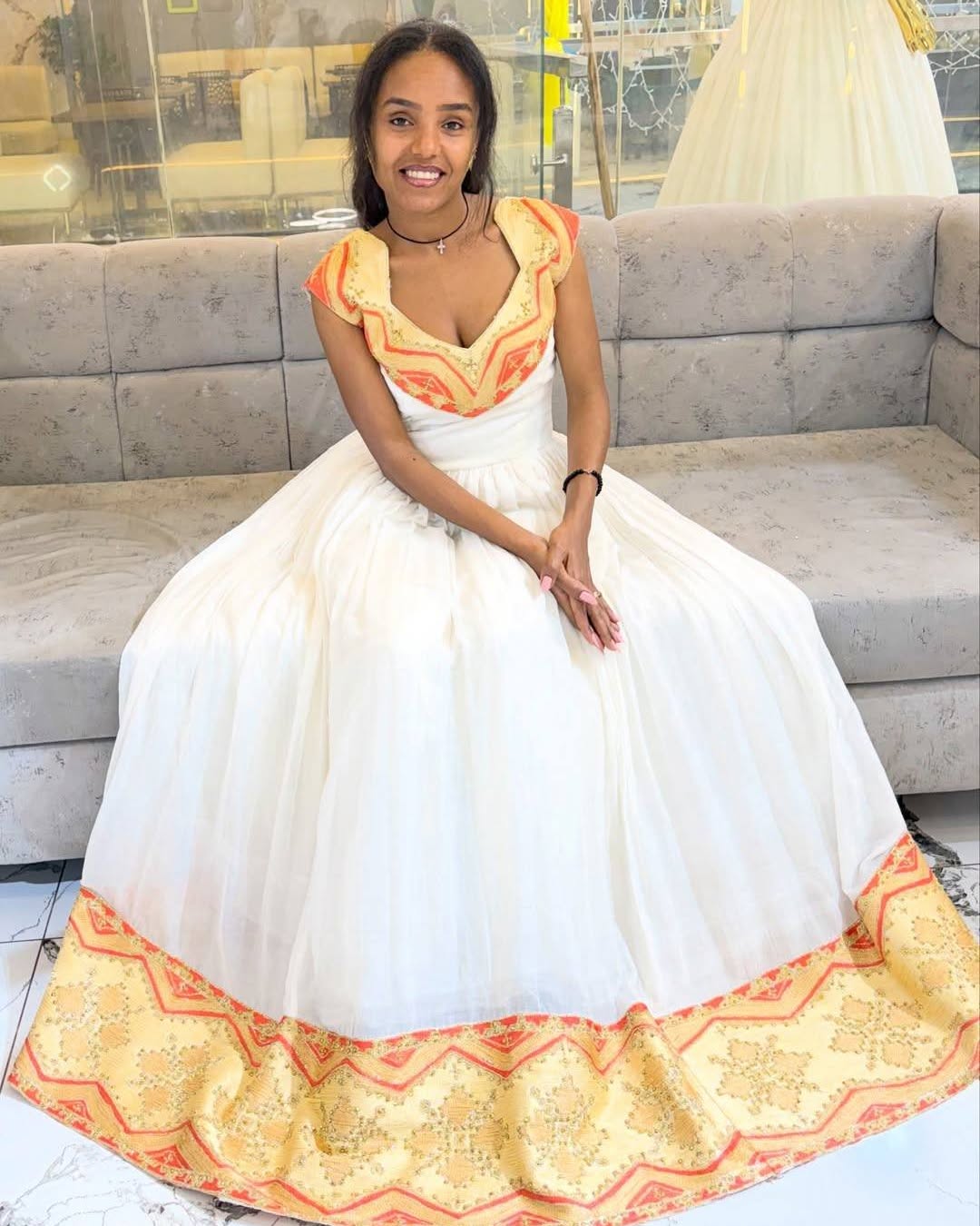 A woman wearing a white ball Habesha Kemis with orange and yellow embroidered details, sitting on a gray sofa in a bridal shop.