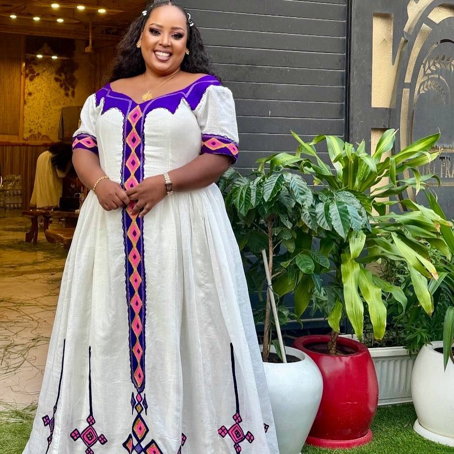 A woman wearing a white A-line Habesha Dress with purple and pink geometric embroidery, standing in an outdoor patio setting with potted plants.