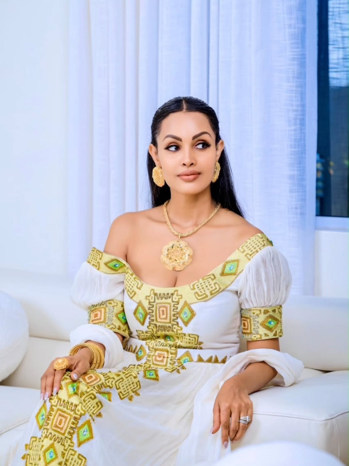 A woman seated on a white sofa wearing a white off-the-shoulder Eritrean Zuria with ornate gold and green geometric embroidery, along with gold jewelry, in a bright indoor setting with white curtains.