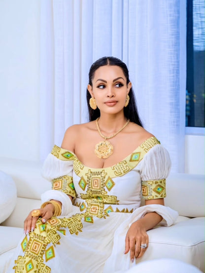 A woman seated on a white sofa wearing a white off-the-shoulder Eritrean Zuria with ornate gold and green geometric embroidery, along with gold jewelry, in a bright indoor setting with white curtains.