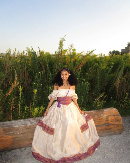 A woman wearing a cream off-the-shoulder bohemian-style Ethiopian National Dresses with purple and orange patterned trim, posing outdoors on a large log in front of green plants.
