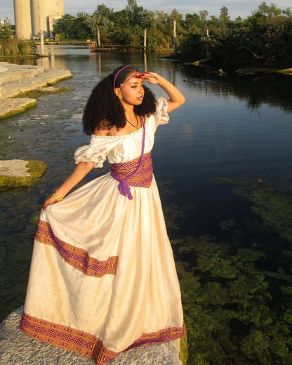 A woman wearing a long cream-colored bohemian-style Ethiopian National Dresses with purple and orange patterned trim, standing on a stone ledge next to a calm body of water at sunset.