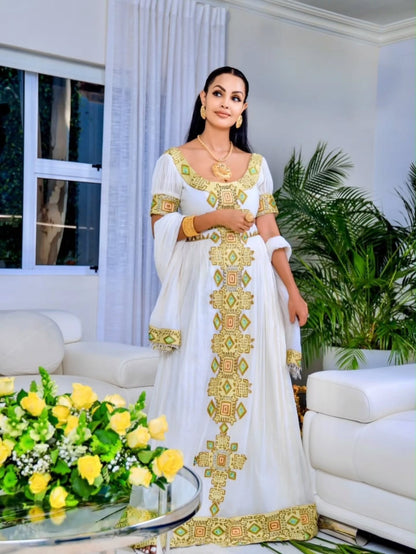 A woman wearing a white traditional Eritrean Zuria with ornate gold and green embroidery, standing in a bright living room with white sofas, green plants, and a glass table with yellow flowers.
