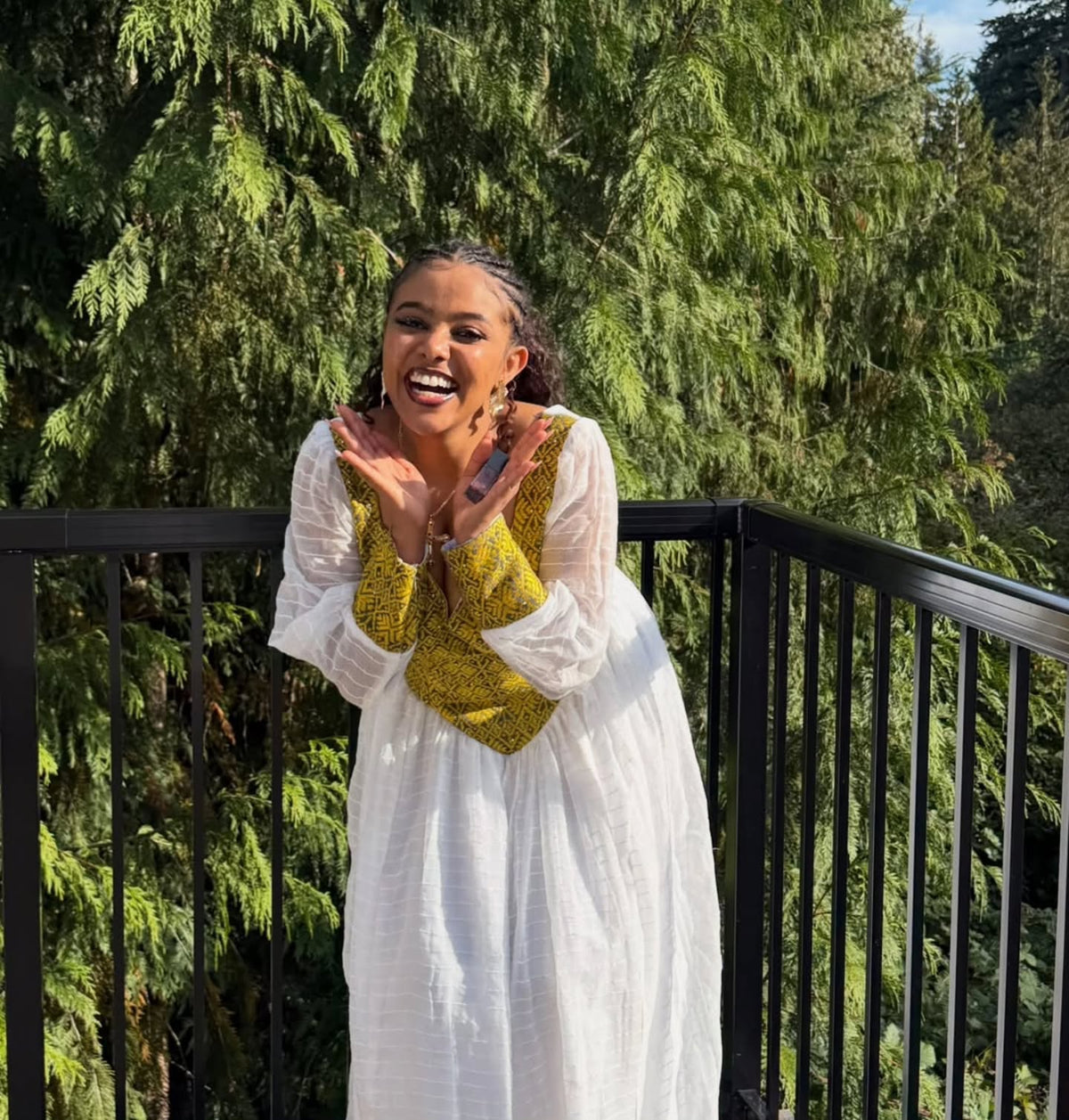 A woman smiling and clapping while wearing a white long-sleeve maxi Habesha Dress with a yellow patterned bow detail, standing behind a black metal railing with green trees in the background.