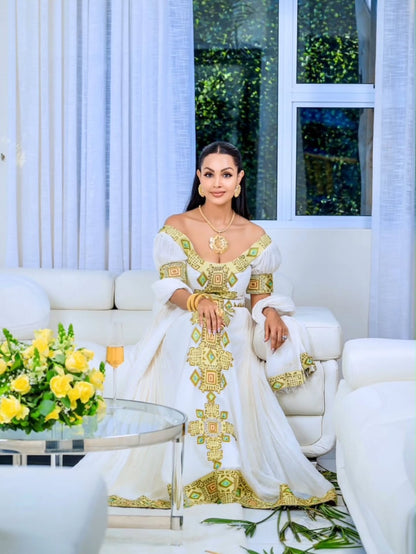 A woman wearing a white Moroccan-style caftan with gold embroidery Eritrean Zuria sits on a white sofa in a bright, modern living room setting.