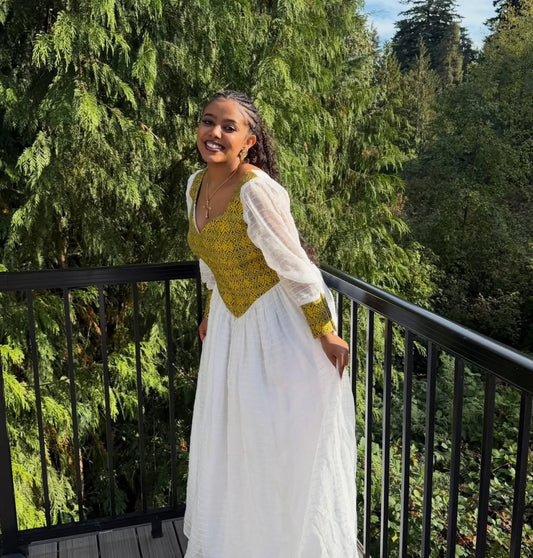 A woman wearing a white Habesha Dress with a yellow embroidered bodice and sheer white puff sleeves, standing on a balcony with a lush green forest background.