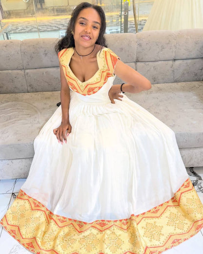 A woman wearing a white A-line Habesha Kemis with orange and yellow patterned embroidery on the neckline and hem, sitting on a gray sofa.