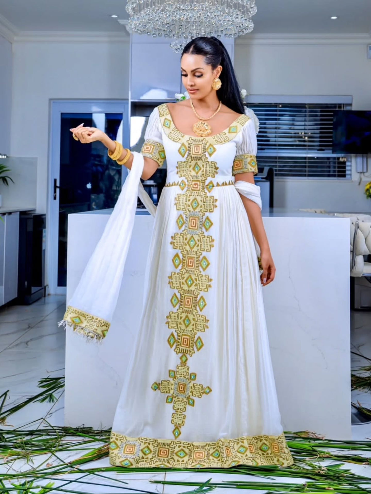A woman models a white traditional long Eritrean Zuria with ornate gold and green embroidery, standing indoors in a modern room with a white display block behind her.
