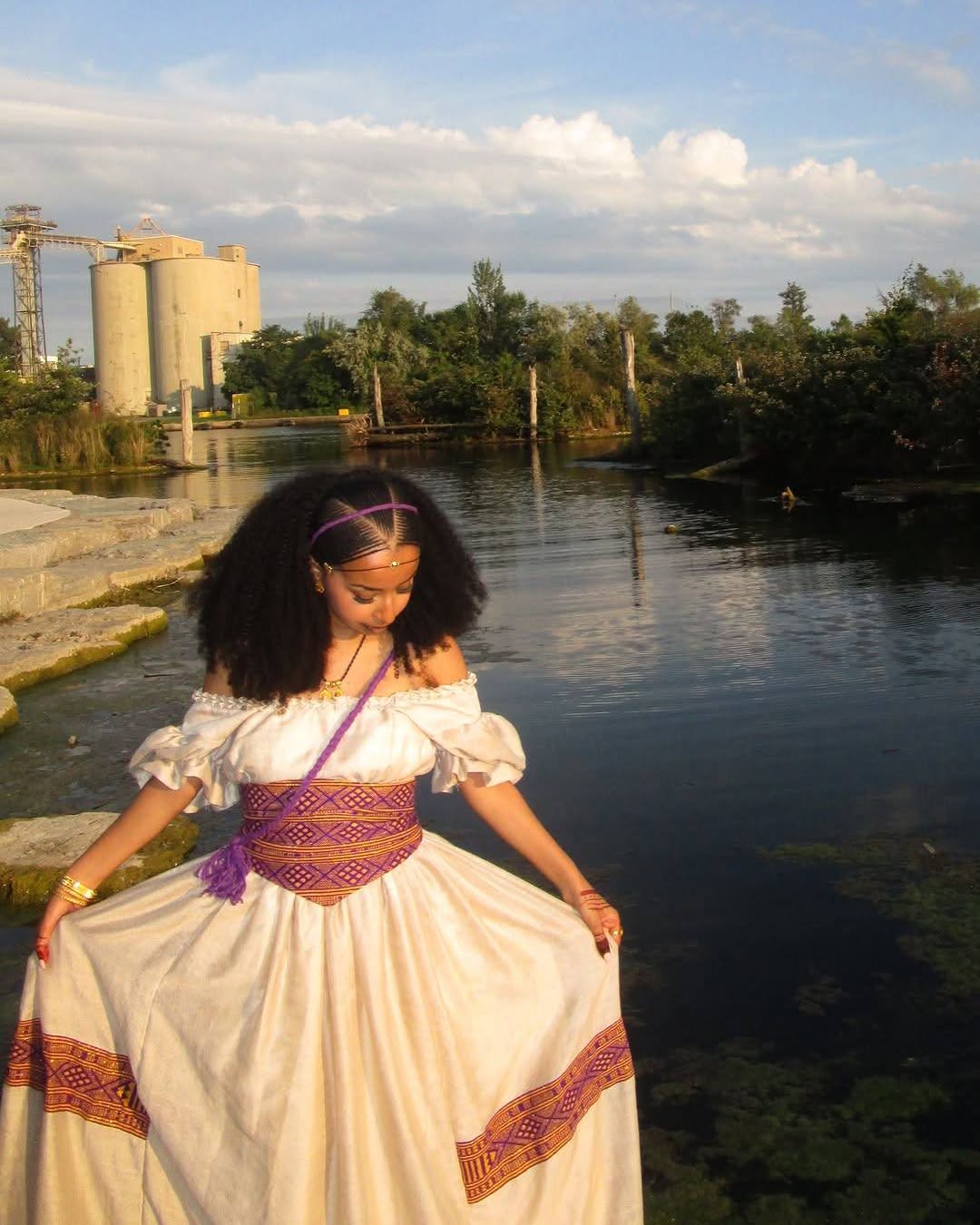 A woman wearing a white off-the-shoulder Ethiopian National Dresses with purple and orange embroidered details stands by a body of water, holding out the sides of the skirt to show its full length. Industrial silos and trees are visible in the background under a cloudy sky.