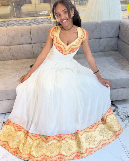 A woman wearing a white A-line Habesha Kemis with orange and yellow embroidered details on the bodice and hem, sitting on a gray sofa.