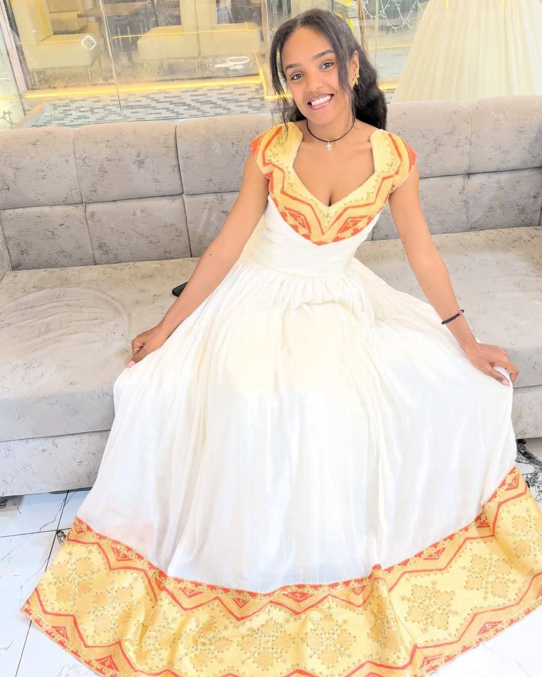A woman wearing a white ball Habesha Kemis with yellow and orange embroidered details, sitting on a gray sofa in a showroom.