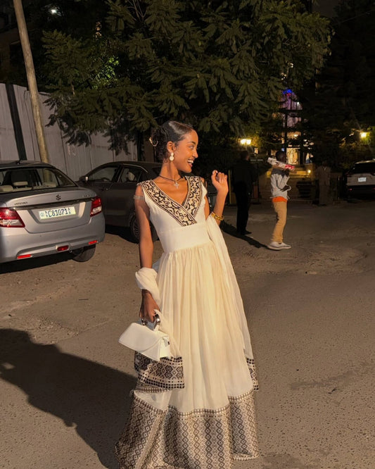 A woman wearing a long cream-colored Ethiopian dress with intricate black and gold embroidery on the bodice and hem, standing outdoors at night on a street with cars and trees in the background.