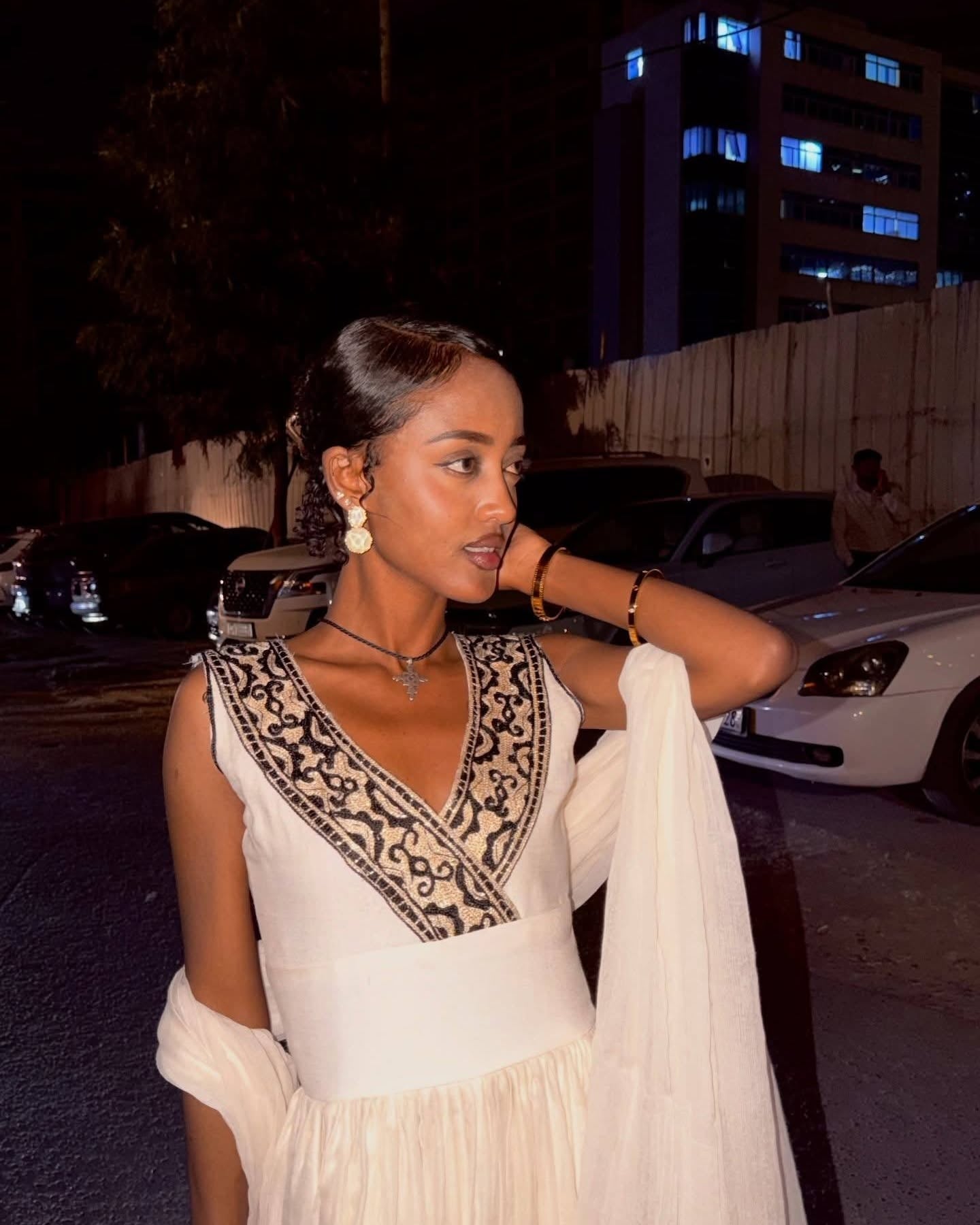 A woman wearing a white sleeveless Ethiopian dress with black embroidered detailing around the V-neckline, photographed outdoors at night with cars and a building in the background.