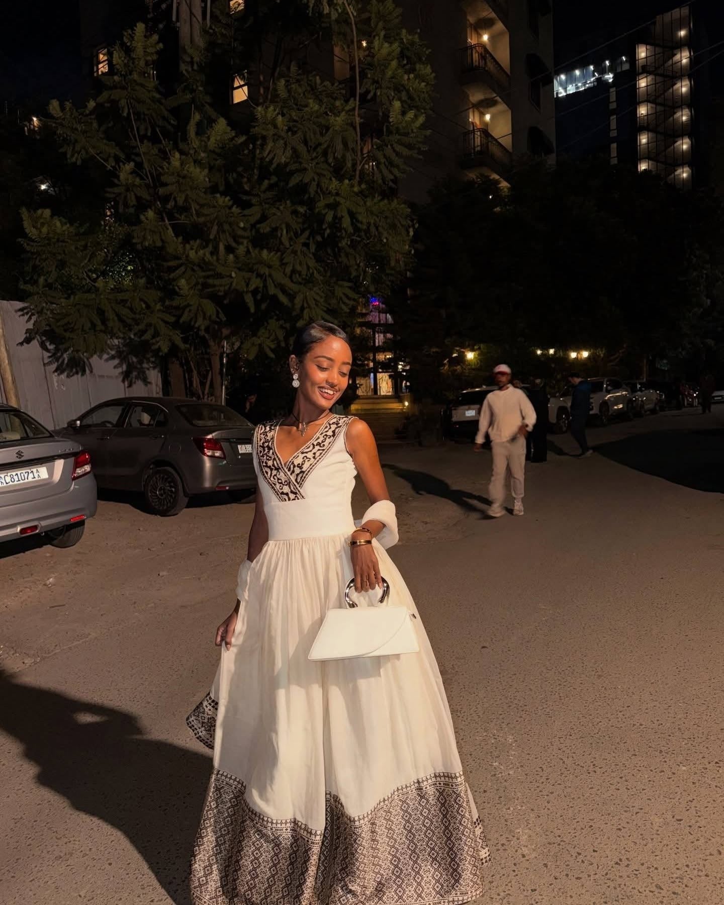 A woman wearing a sleeveless white maxi Ethiopian dress with a patterned neckline and a sheer layered skirt, holding a white handbag while walking outdoors at night on a street with cars and trees in the background.