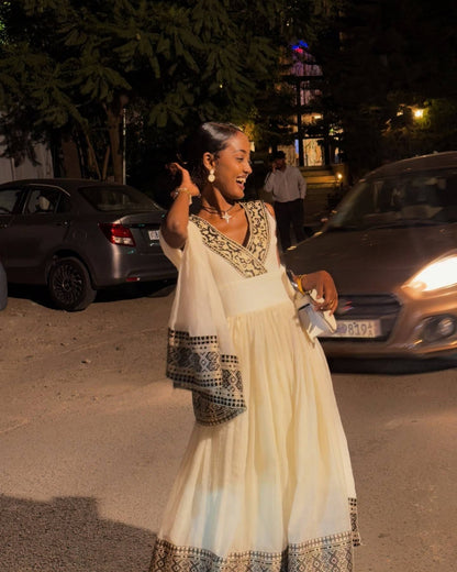 A woman wearing a cream-colored Ethiopian dress with black and gold embroidery, standing outdoors at night near parked cars.