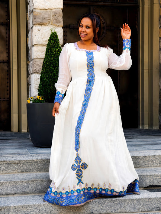 A woman wearing a long white Habesha Store  with blue embroidered details stands on stone steps in front of a building entrance. The dress has sheer long sleeves, a high neckline, and a vertical and horizontal blue embroidered pattern on the front and cuffs.