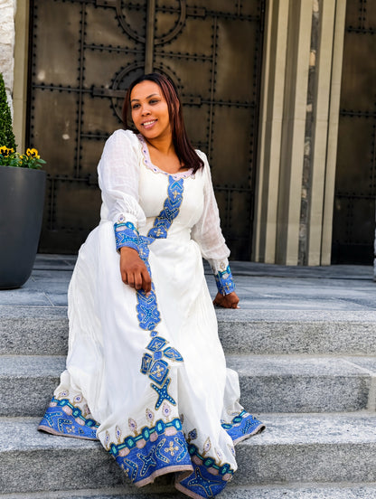 A woman wearing a white long-sleeve maxi Habesha Store  with blue and turquoise geometric embroidery sits on stone steps in front of a large metal door.